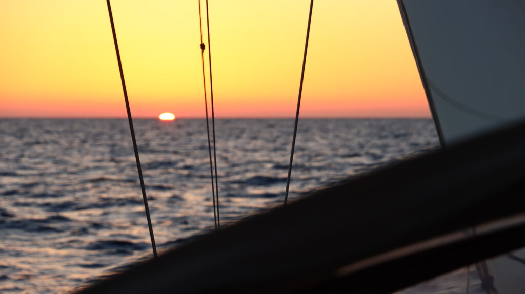 Sunset over open ocean framed by sailboat rigging during a passage